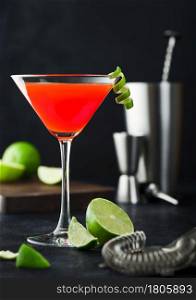 Cosmopolitan cocktail in classic crystal glass with lime peel and fresh limes with strainer, jigger and shaker on black table background.