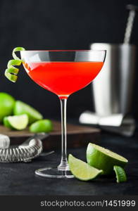 Cosmopolitan cocktail in luxury crystal glass with lime peel and fresh limes with strainer, jigger and shaker on black table background.