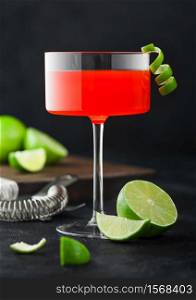 Cosmopolitan cocktail in modern crystal glass with lime peel and fresh limes with strainer on black table background.