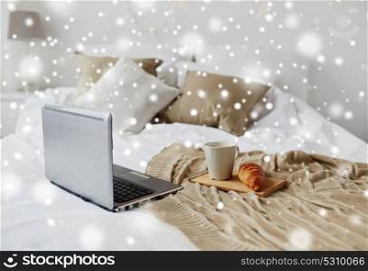 cosy home, interior and winter concept - cozy bedroom with laptop computer, coffee cup and croissant on bed at home over snow. laptop, coffee and croissant on bed at cozy home
