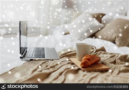 cosy home, interior and winter concept - cozy bedroom with laptop computer, coffee cup and croissant on bed over snow. laptop, coffee and croissant on bed at cozy home