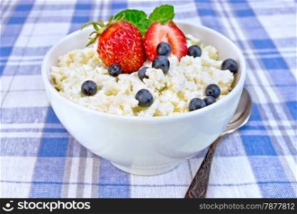 Cottage cheese in a white bowl with strawberries, blueberries and mint, a spoon against a background of blue linen tablecloth