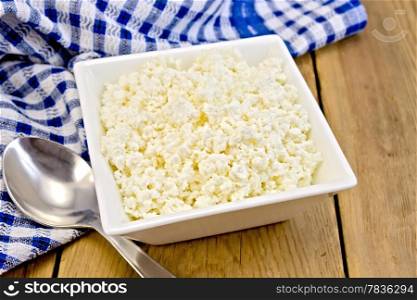 Cottage cheese in a white square bowl, blue checkered napkin, spoon on a wooden board