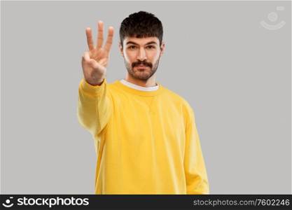 count and people concept - young man in yellow sweatshirt showing three fingers over grey background. man in yellow sweatshirt showing three fingers