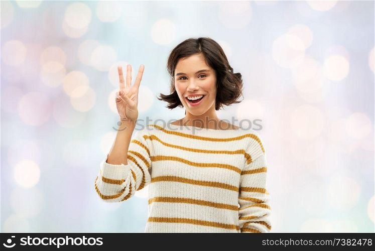 counting and people concept - happy smiling young woman in striped pullover showing three fingers over festive lights background. happy smiling woman showing three fingers