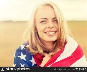 country, patriotism, independence day and people concept - happy smiling young woman wrapped into national american flag on cereal field. happy woman in american flag on cereal field