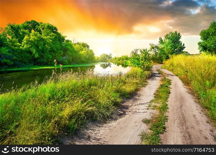 Country road to the sun along the river at sunset. Green vibrant trees. Summer landscape. Country road to sun along river at sunset