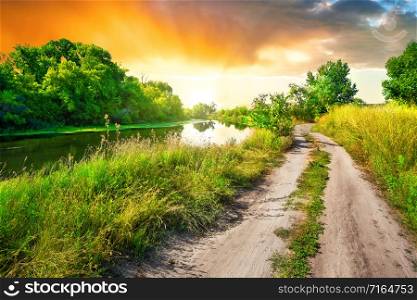 Country road to the sun along the river at sunset. Green vibrant trees. Summer landscape. Country road to sun along river at sunset