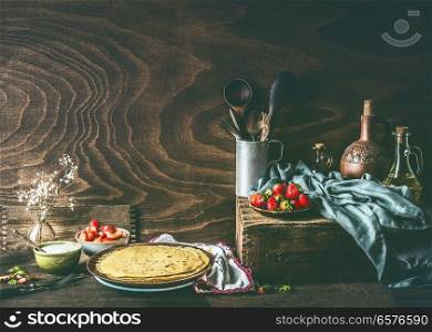Country still life with homemade crepes on dark rustic wooden kitchen table with strawberries and yogurt in bowls .