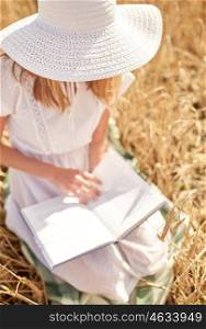 country, summer holidays, literature and people concept - close up of young woman in white straw hat and dress reading book on cereal field