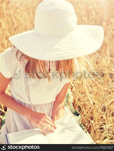 country, summer holidays, literature and people concept - close up of young woman in white straw hat and dress reading book on cereal field. close up of woman reading book on cereal field