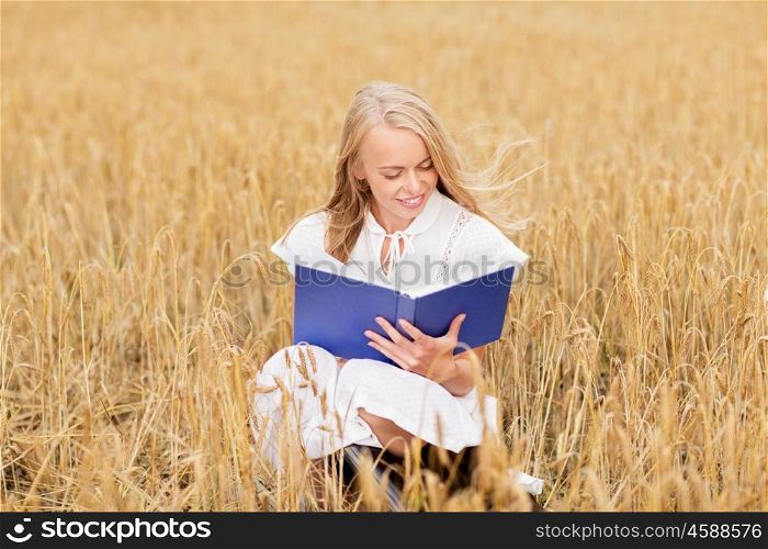 country, summer holidays, literature and people concept - smiling young woman in white dress reading book on cereal field