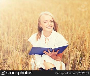 country, summer holidays, literature and people concept - smiling young woman in white dress reading book on cereal field. smiling young woman reading book on cereal field