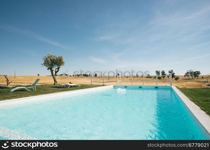 Country Tourism Lounge Pool Landscape. A relaxing country scene in Alentejo, Portugal, with a fantastic landscape view over a big, blue swimming pool. Also Includes two lounge chairs and a small olive tree.