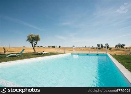 Country Tourism Lounge Pool Landscape. A relaxing country scene in Alentejo, Portugal, with a fantastic landscape view over a big, blue swimming pool. Also Includes two lounge chairs and a small olive tree.