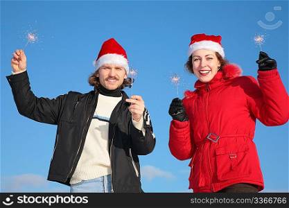 couple against blue sky background in winter in santa claus hats with sparklers