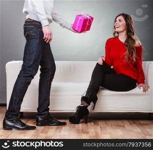 Couple and holiday concept. man surprising cheerful woman with a gift box, girl sitting on couch at home