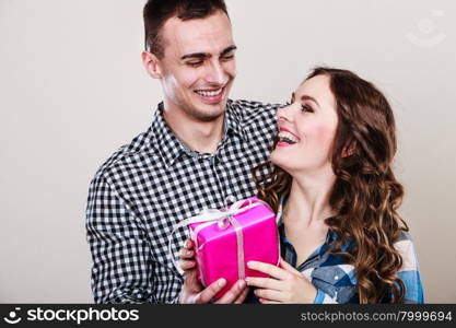 Couple and holiday concept. Smiling young man surprising cheerful woman with pink gift box on gray background