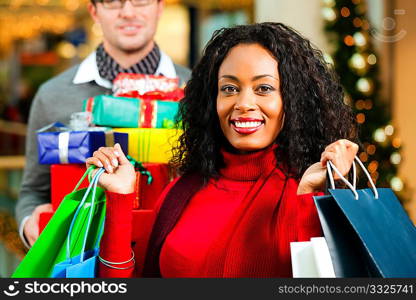 Couple - Caucasian man and black woman - with Christmas presents, gifts and shopping bags - in a mall in front of a Christmas tree