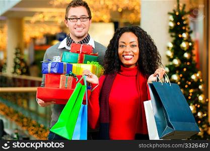 Couple - Caucasian man and black woman - with Christmas presents, gifts and shopping bags - in a mall in front of a Christmas tree