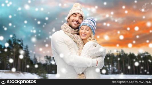 couple, christmas and holidays concept - smiling man and woman in hats and scarf hugging over winter forest background. smiling couple in winter clothes hugging
