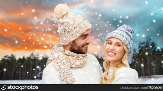 couple, christmas and holidays concept - smiling man and woman in hats and scarf over winter forest background. smiling couple in winter clothes hugging