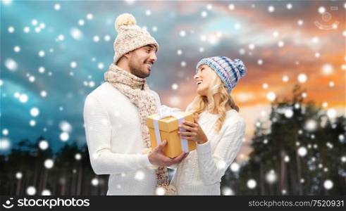 couple, christmas and holidays concept - smiling man and woman in hats and scarf with gift box over winter forest background. smiling couple in winter clothes with gift box
