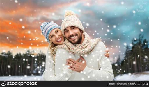 couple, christmas and holidays concept - smiling man and woman in hats and scarf hugging over winter forest background. smiling couple in winter clothes hugging
