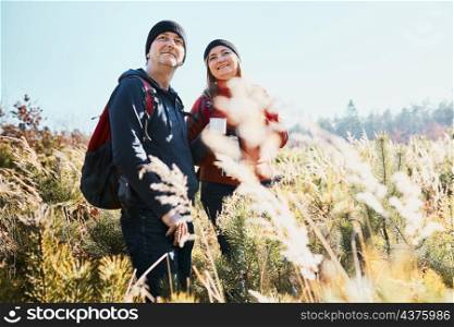 Couple enjoying nature while vacation trip. Hikers with backpacks looking at mountains view. People standing in tall grass on path to mountains on sunny day. Active leisure time close to nature