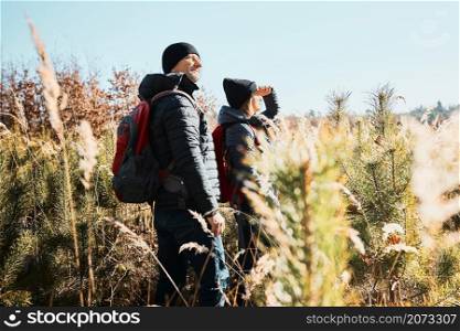 Couple exploring nature while vacation trip. Hikers with backpacks on way to mountains. People walking through tall grass along path in meadow on sunny day. Active leisure time close to nature
