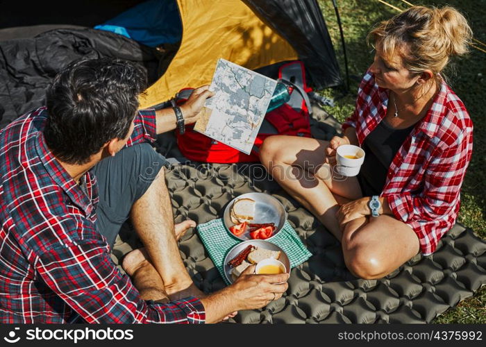 Couple having breakfast and planning their day sitting by tent at camping. People actively spending summer vacations close to nature outdoors. Concept of camp life