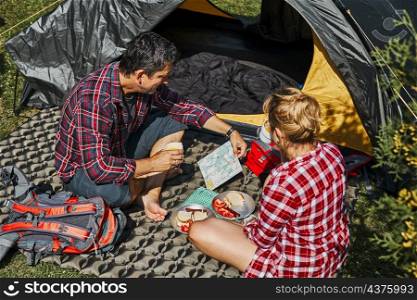 Couple having breakfast and planning their day sitting by tent at camping. People actively spending summer vacations close to nature outdoors. Concept of camp life