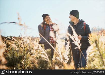 Couple having fun while vacation trip. Hikers with backpacks on way to mountains. People exploring nature walking through tall grass along path in meadow on sunny day. Active leisure time close to nature