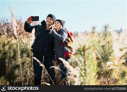 Couple having video call sending greetings from vacation trip. Hikers with backpacks on way to mountains. People walking through tall grass along path in meadow on sunny day. Active leisure time close to nature