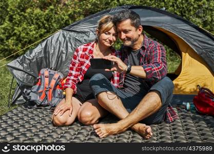 Couple having video call with friends using smartphone while sitting in tent at camping. People relaxing in tent during summer vacation. Actively spending vacations outdoors close to nature. Concept of camp life