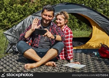 Couple having video call with friends using smartphone while sitting in tent at camping. People relaxing in tent during summer vacation. Actively spending vacations outdoors close to nature. Concept of camp life