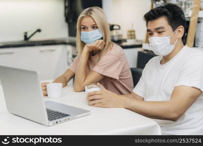 couple home with medical masks during pandemic working laptop