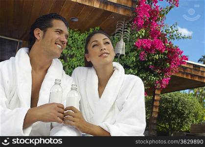 Couple in bathrobes holding water bottles and looking away on porch