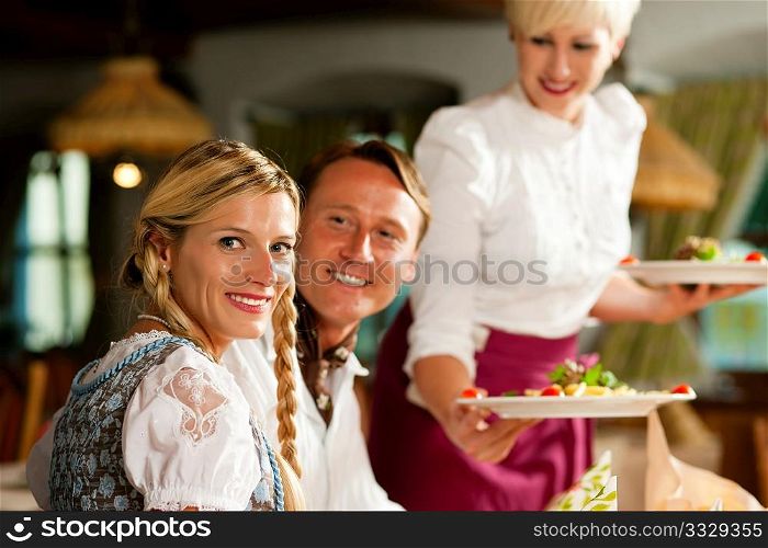 Couple in Bavarian Tracht drinking wheat beer in a typical pub, the waitress is serving the drinks