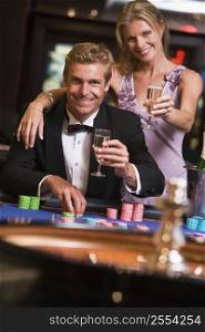 Couple in casino at roulette table holding up champagne and smiling (selective focus)
