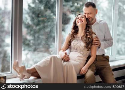 couple in love in festive clothes sitting and hugging near the large window and Christmas tree in decorated studio. happy holiday in winter time.. couple in love in festive clothes sitting and hugging near the large window and Christmas tree in decorated studio. happy holiday in winter time