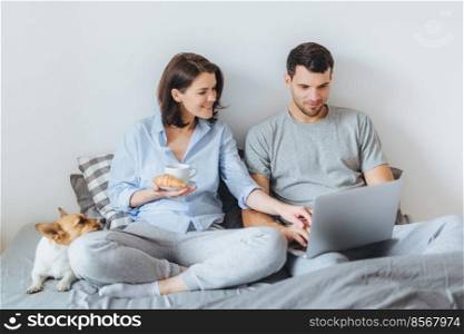 Couple in love spend morning weekends in bedroom, view photos on laptop computer, sit on bed together with their pet, being in good mood. Female drinks coffee with crroissant sits near husband