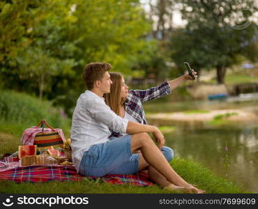 Couple in love taking a selfie by mobile phone while enjoying picnic time drink and food in beautiful nature on the river bank