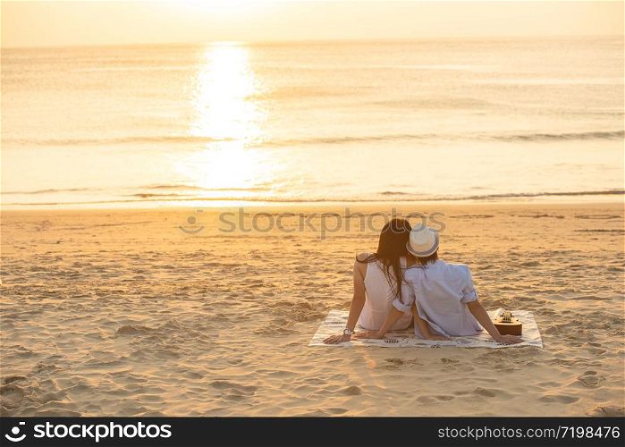 Couple in love watching sunset together on beach travel summer holidays. People silhouette from behind sitting enjoying view sunset sea on tropical destination vacation. Romantic couple on the beach.