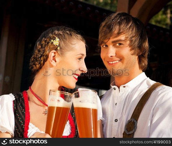 Couple in traditional Bavarian Tracht - Dirndl and Lederhosen - in front of a beer tent at the Oktoberfest or in a beer garden enjoying a glass of tasty wheat beer