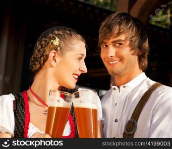 Couple in traditional Bavarian Tracht - Dirndl and Lederhosen - in front of a beer tent at the Oktoberfest or in a beer garden enjoying a glass of tasty wheat beer
