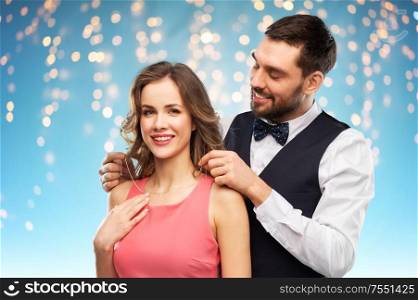 couple, jewelry and people concept - happy man puts necklace with diamond pendant on his girlfriend over holiday lights on blue background. happy man puts necklace on his girlfriend