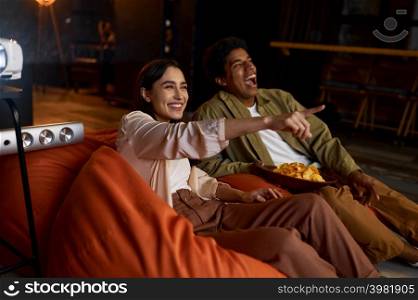 Couple laughing pointing on screen. Family watching television projector on bag seat in living room. Family couple watching comedian film on projector