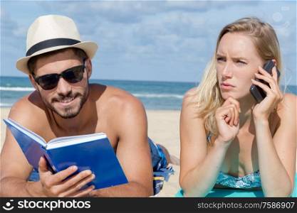 couple layed on beach man readying book woman on cellphone