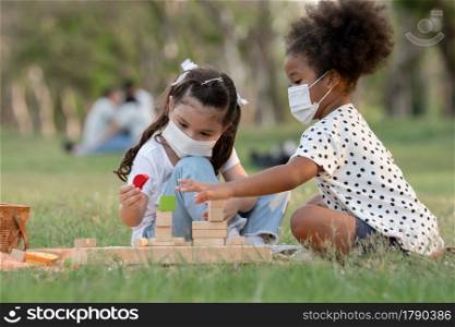 Couple little girls African and Caucasian kids wear face mask while sitting and playing wooden blocks toy in green park together. Ethnic diversity of friendship in new normal lifestyle concept